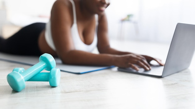 Dodue jeune femme noire allongée sur un tapis de yoga et regardant une vidéo de sport en ligne sur un ordinateur portable à la maison