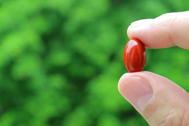 Les doigts de l'homme tenant une pilule de supplément de gélule rouge contre le feuillage vert flou