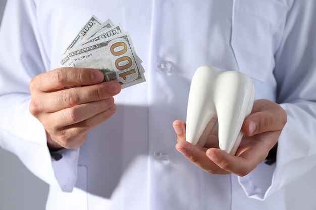 Des dollars et une dent dans les mains d'un dentiste sur un fond gris.