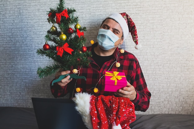 Don de cadeaux de Noël. Homme en masque médical partageant des cadeaux par le biais d'appels vidéo. Vacances dans l'isolement. Arbre du nouvel an et lumières avec décorations. Famille et amis célébrant en ligne.