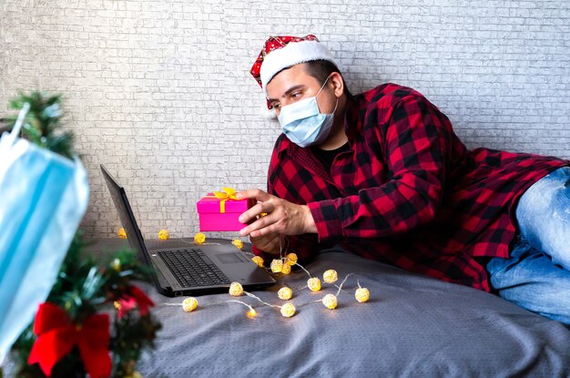 Don de cadeaux de Noël. Homme en masque médical partageant des cadeaux par le biais d'appels vidéo. Vacances dans l'isolement. Arbre du nouvel an et lumières avec décorations. Famille et amis célébrant en ligne.