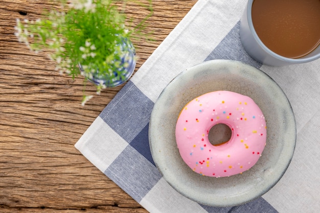 Donut aux fraises rose dans une assiette en céramique gris clair à côté du café et du vase sur serviette et bois rustique