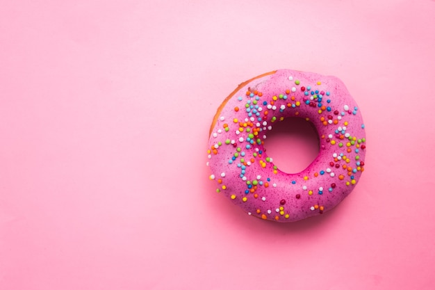 Donut circulaire fait maison avec glaçage rose et pépites d'arc-en-ciel sur le rose tendre tendance situé sur le côté droit.