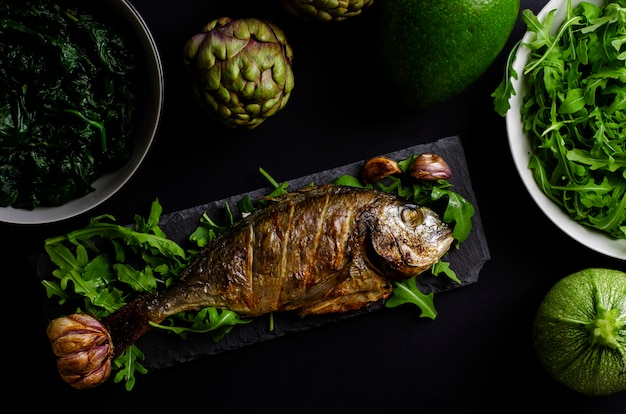 Dorade Cuite Au Four Sur Une Assiette De Service Avec De La Roquette Et Des Légumes Verts Sur Fond Noir