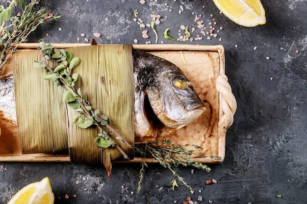 Photo dorade grillée fraîche éviscérée ou poisson dorado servi avec citron, salade verte, romarin, huile d'olive, vin blanc et sel rose sur une assiette en céramique enveloppée de feuilles de bambou sur une surface sombre.