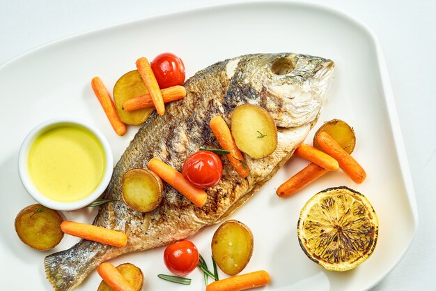 Dorado grillé avec pommes de terre, carottes et tomates cerises dans une assiette blanche. Isolé sur une surface blanche. Vue d'en-haut