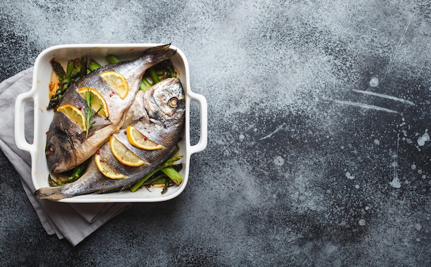 Dorado de poisson au four avec asperges vertes dans un plat de cuisson en céramique blanche sur fond de béton rustique gris, vue de dessus. Dîner sain avec concept de poisson, régime et alimentation saine. Espace pour le texte