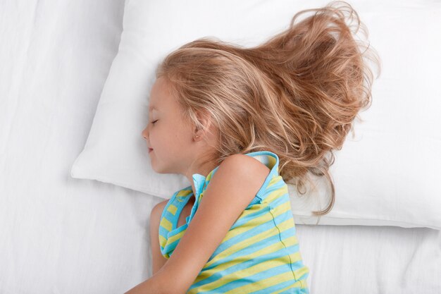 Dormir jolie fille aux cheveux clairs se trouve sur le côté dans un lit confortable, vêtue de vêtements de nuit, a des rêves agréables, pose sur des draps blancs