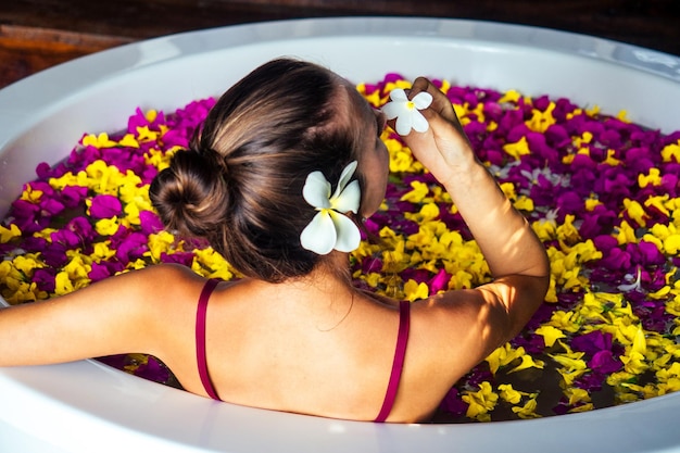 Dos d'une femme dans une baignoire avec des fleurs exotiques tropicales dans l'eau et une fleur de plumeria blanche dans ses cheveux. fille dans un hôtel écologique prenant un bain dans le salon spa vacances d'été dans une chambre de luxe, soins de la peau