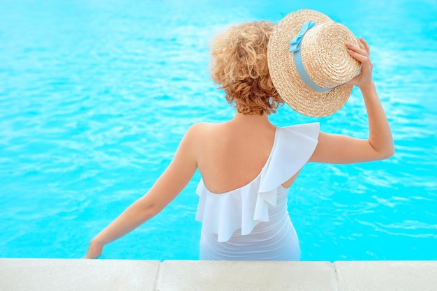 le dos de la femme rousse dans le maillot de bain blanc assis au bord de la piscine dans le chapeau de paille