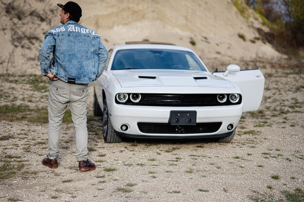 Dos d'homme en veste de jeans et casquette près de sa voiture de muscle blanche en carrière à Los Angeles