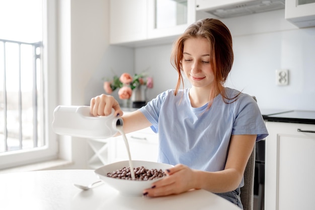 Une douce adolescente verse du lait dans un bol avec un petit déjeuner sec dans la cuisine.