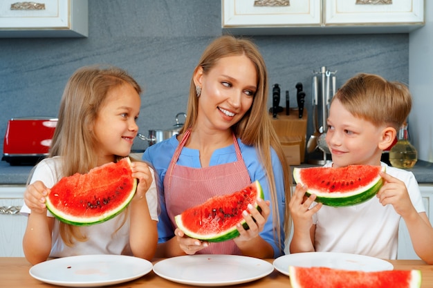 Douce famille, mère et ses enfants mangeant de la pastèque dans leur cuisine s'amusant