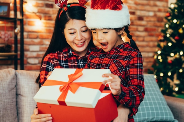 douce famille ouvrant une grande boîte cadeau le lendemain de Noël reçue la veille de noël la nuit. deux filles de noël célébrant les vacances dans une chambre décorée dans une maison chaleureuse. ruban arc rouge sur blanc présent.