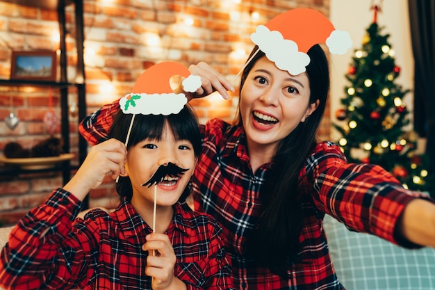 douce famille s'amusant à jouer avec une décoration en papier faisant un drôle de visage prenant un selfie à la maison pour célébrer la veille de noël. les jolies filles du père noël aiment passer du temps ensemble en vacances.