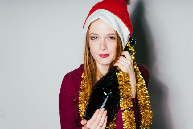 Une douce fille rousse dans un bonnet rouge comme le Père Noël célèbre la nouvelle année, tient une bouteille de champagne