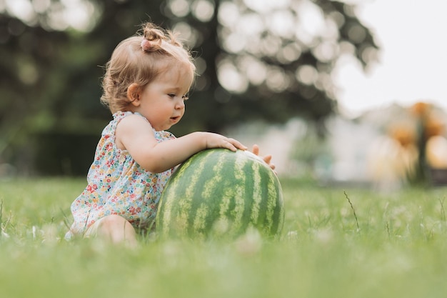 douce petite fille avec une énorme pastèque est assise sur une pelouse verte enfant joue à l'extérieur style de vie