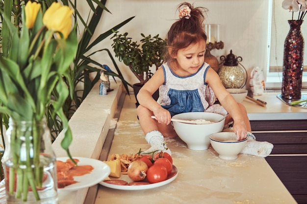 Douce petite fille mignonne apprend à cuisiner un repas dans la cuisine tout en étant assise sur un comptoir.