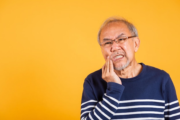 Douleur dentaire Asiatique homme âgé malheureux problèmes avec la gencive douleur studio tourné isolé sur fond jaune Portrait de vieil homme âgé triste main touchant la joue souffrant de maux de dents soins dentaires