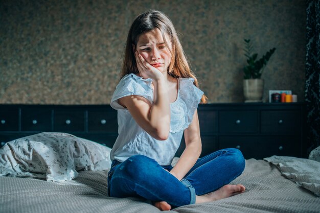 Douleur dentaire. Belle fille ressentant une forte douleur, maux de dents. Une fille est assise sur un lit dans sa chambre et met une main sur sa joue.