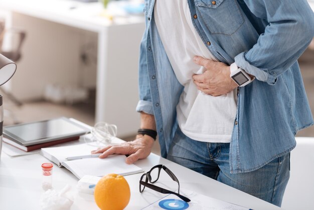 Douleur insupportable. Homme malade portant des vêtements décontractés mettant la main gauche sur l'estomac en se tenant debout derrière le lieu de travail