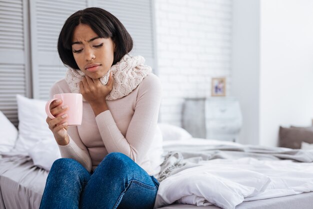 Douloureux. Jeune femme fatiguée malheureuse toucher son cou tout en buvant du thé chaud et souffrant de maux de gorge