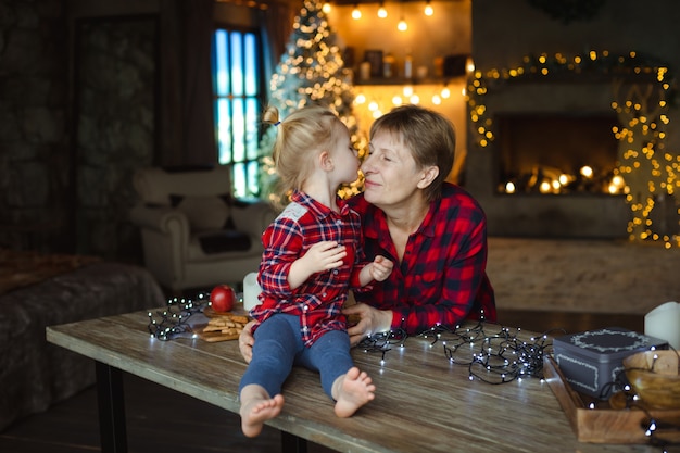 Un Doux Bambin Embrasse Sa Grand-mère Dans Le Nez, Assis Sur Une Table En Bois Dans Une Maison De Chasse Décorée Pour Noël.