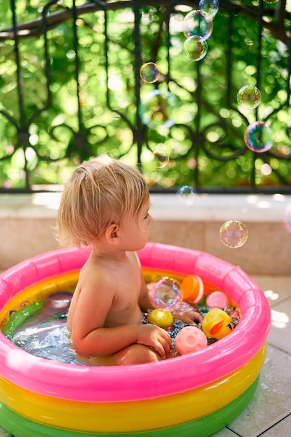 Doux petit bébé est assis dans une petite piscine gonflable avec vue latérale sur les jouets