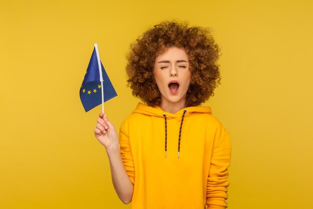 Photo droit de vote civil, droit de l'ue. portrait d'une jeune femme aux cheveux bouclés en sweat à capuche tenant le drapeau de l'union européenne et gardant la bouche grande ouverte, déclarant haut et fort ses droits. tourné en studio intérieur, isolé