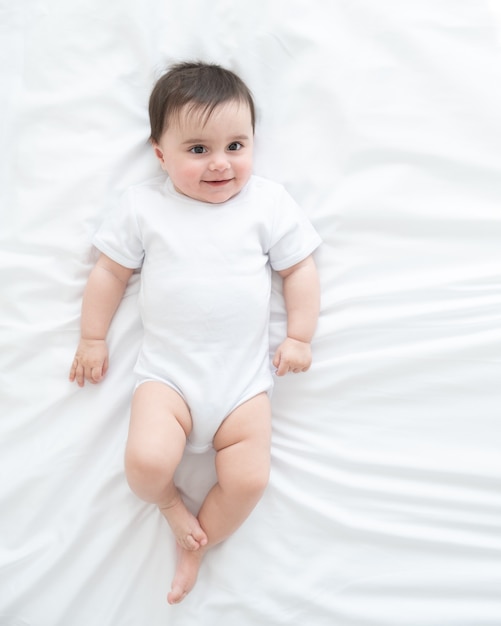 Drôle bébé en bonne santé 6 mois en body blanc allongé sur le lit sur une literie blanche. vue de dessus.