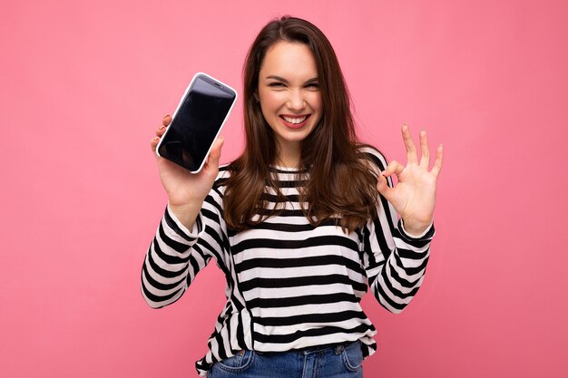 Drôle Belle Jeune Femme Heureuse Portant Un Pull Rayé Isolé Sur Fond Avec Copie Espace Montrant Le Geste Ok Regardant La Caméra Montrant L'écran Du Téléphone Mobile. Maquette, Découpe