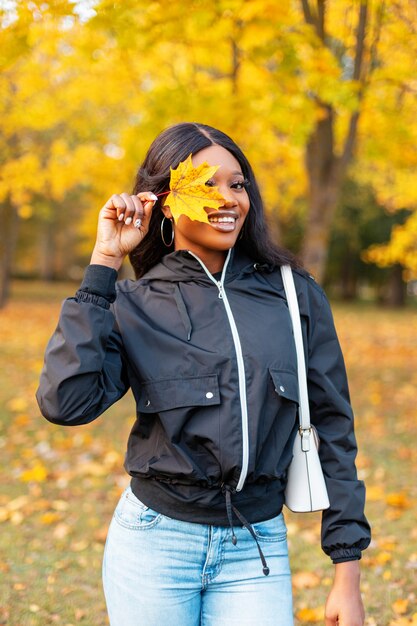 Drôle belle jeune fille afro-américaine avec un sourire dans des vêtements décontractés à la mode couvre ses yeux avec une feuille jaune et se promène dans le parc avec un feuillage d'automne doré brillant