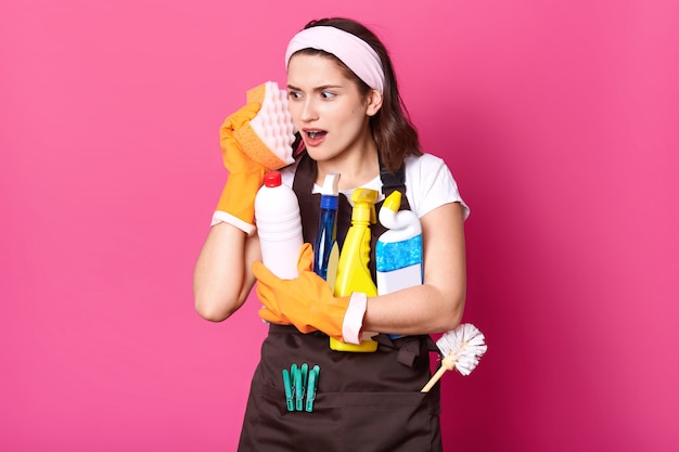 Drôle charmante jeune femme au foyer caucasienne portant un t-shirt décontracté et un tablier, des gants en caoutchouc orange, tenant des éponges sur son visage, imagine que c'est un téléphone, s'amuse tout en faisant des tâches. Concept d'hygiène.