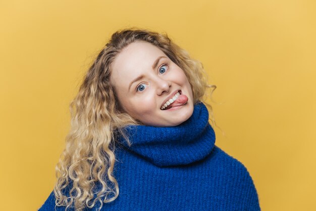 Drôle comique frisée jeune femme avec une expression positive, montre la langue, s'amuse avec des amis