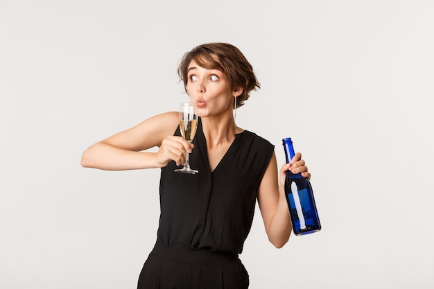 Drôle femme ivre faire la fête, tenant une bouteille et siroter du champagne dans un verre, debout sur blanc