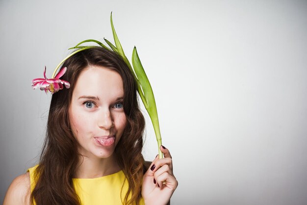 Une drôle de fille aux yeux bleus en robe jaune montre la langue et tient une fleur