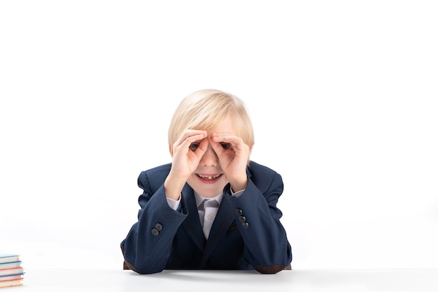 Drôle garçon blond en uniforme scolaire a fait ses mains comme des jumelles Portrait d'écolier s'amuser est isolé sur fond blanc