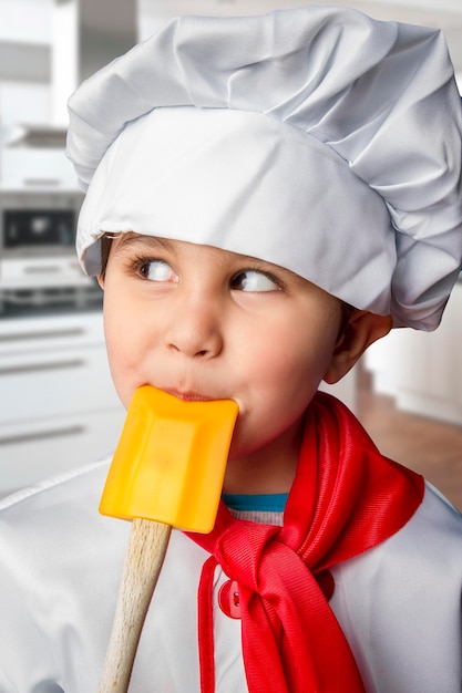 Drôle de garçon habillé en chef avec une cuillère orange dans la bouche