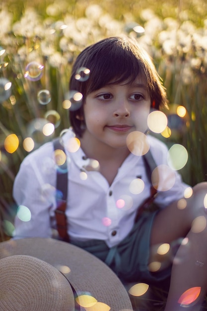 Drôle heureux un beau garçon enfant assis dans un chapeau sur un champ avec des pissenlits blancs au coucher du soleil en été des bulles de savon volent