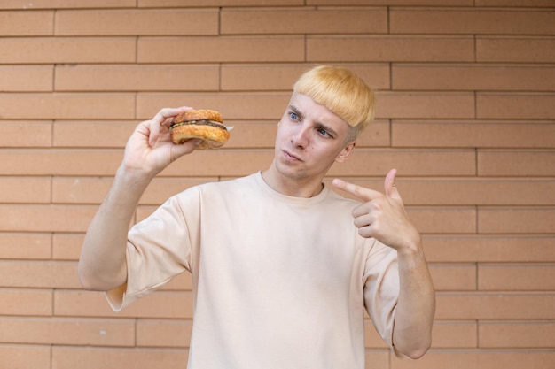 Un drôle d'homme européen blond vêtu d'un t-shirt beige tenant un hamburger pointant et le regardant