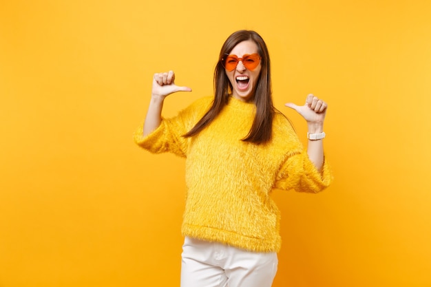 Drôle de jeune femme folle en pull de fourrure et lunettes orange coeur criant des pouces pointant sur elle-même isolé sur fond jaune vif. Les gens émotions sincères, concept de style de vie. Espace publicitaire.