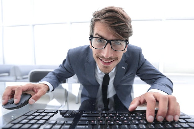 Drôle de jeune homme au visage fou assis à la table avec les mains sur la souris et le clavier de l'ordinateur