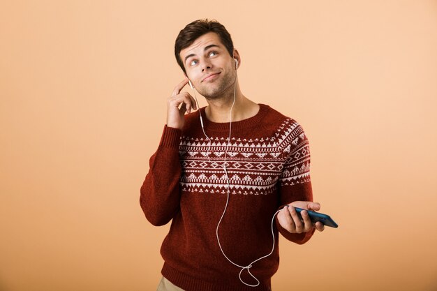 Drôle jeune homme isolé sur mur beige à l'aide de téléphone écoute de la musique par des écouteurs