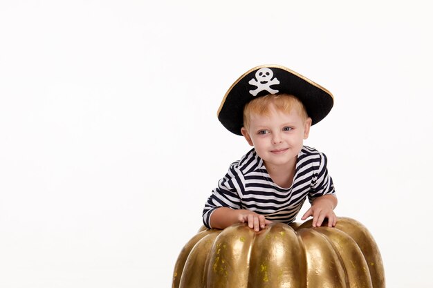 Drôle petit garçon en costumes de carnaval de pirates debout avec une grosse citrouille sur blanc