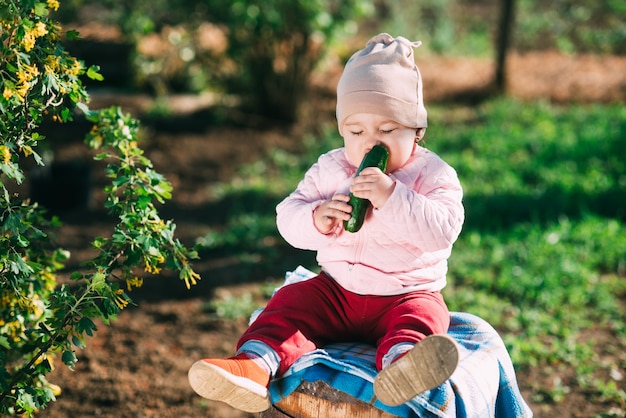 Drôle de petite fille assise sur un tonneau de manger un concombre frais dans le jardin