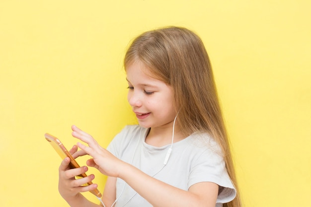 Une drôle de petite fille aux cheveux longs sur fond jaune tient un smartphone dans les mains avec des écouteurs et des sourires. Le concept d'enfants et de gadgets, de réseaux sociaux, de tik tok.