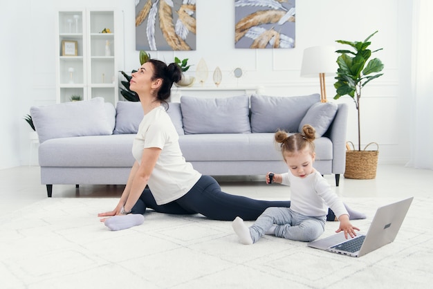 Drôle petite fille jouant avec un ordinateur portable pendant que sa maman saine et sportive a une formation de yoga en ligne à la maison.