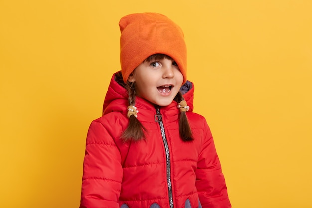 Photo drôle de petite fille posant isolé sur mur jaune avec la bouche largement ouverte, tirez sa casquette sur un œil