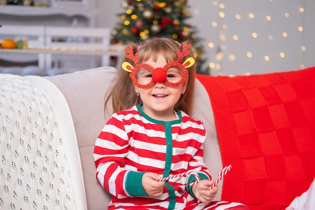 Drôle de petite fille en pyjama de Noël souriante assise sur un canapé profitant de Noël