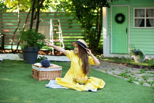 Drôle de petite fille en robe jaune et chapeau de paille assis avec panier pique-nique et fruits sur pelouse